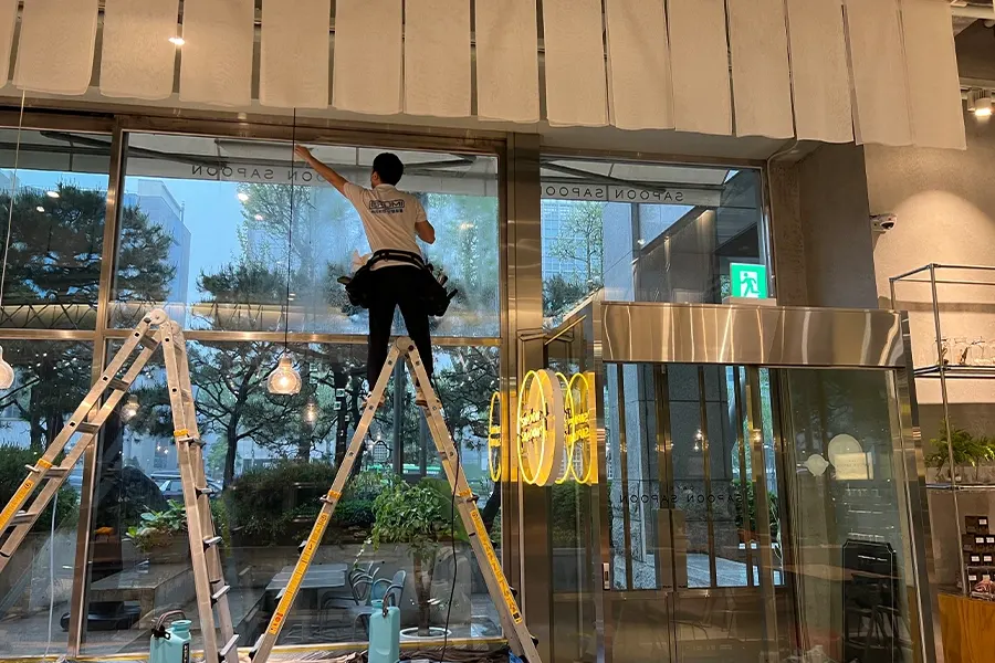 작업자가 사다리 위에서 통유리 상단 모서리까지 필름을 꼼꼼히 부착하는 모습