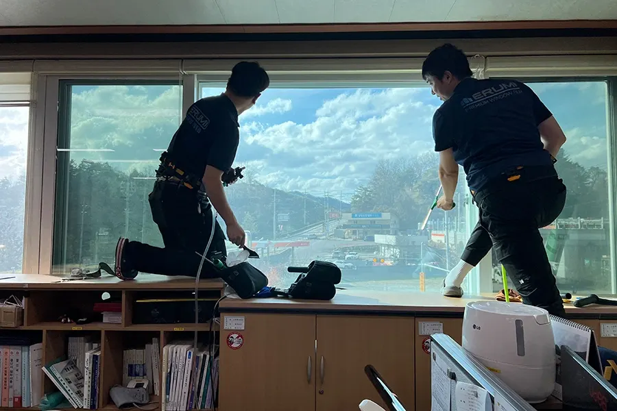 시공 중인 모습, 두 명의 작업자가 창문에 물을 분사하며 필름을 밀착시키는 장면